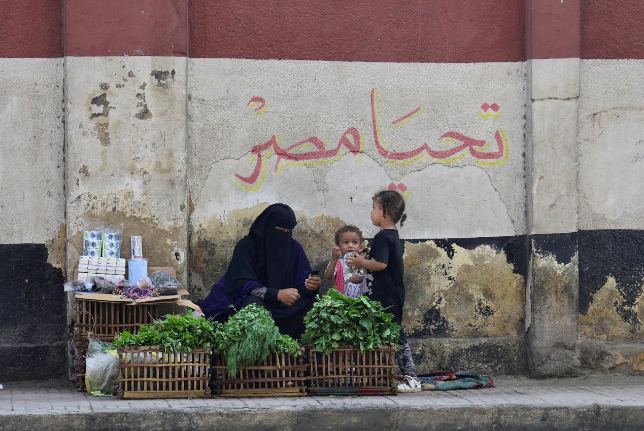 2023年10月10日,埃及開羅,一名帶著孩子的街頭小販坐在一幅壁畫前,壁畫上有埃及國旗和“埃及萬歲”的文字。